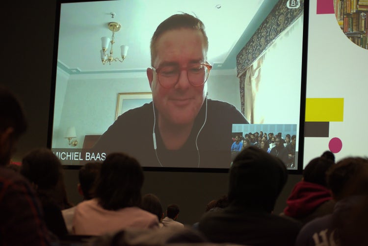 A photograph of a computer conference call screen on which a man with short grey/brown hair, wearing glasses and wired ear buds, is speaking. Audience members are in the foreground.