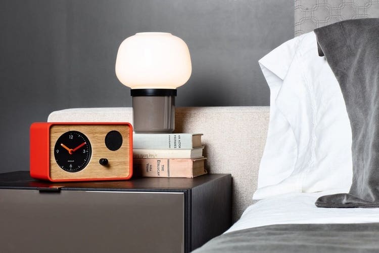 A photograph of a bedside scene featuring a modern table lamp with a soft white glow, stacked books, and a red‑framed wooden clock radio on a dark nightstand. Crisp white bedding and gray pillows create a calm, minimalist bedroom atmosphere.