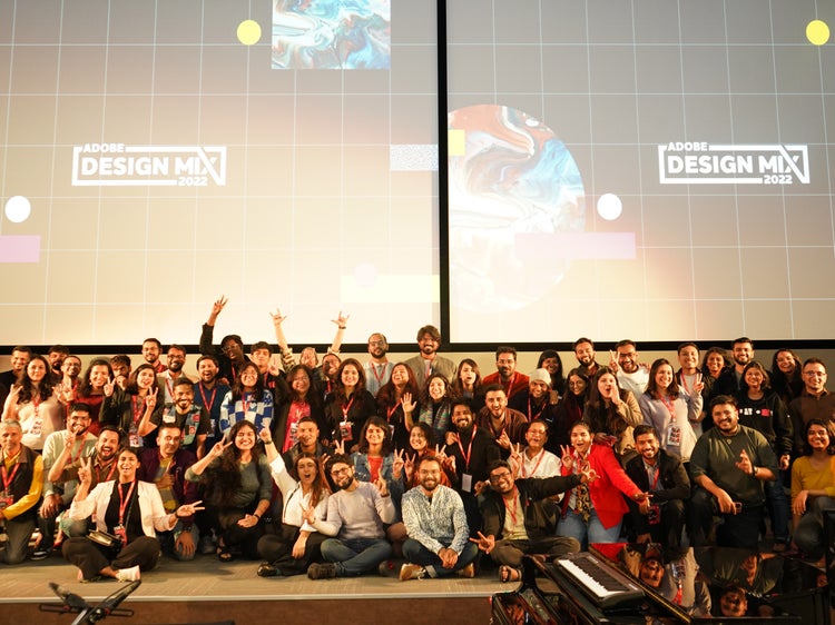 A photograph of about 50 people sitting on a raised platform with the Design Mix logotype in white on a black background projected on two screens behind them.