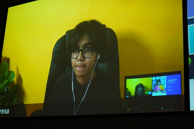 A photograph of a computer conference call screen on which a teenager with short dark hair, wearing a black sweater, glasses, and wired ear buds, is speaking.