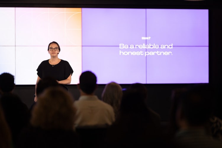 A white woman with dark brown hair, wearing black-rimmed glasses, a black top, and grey slacks stands speaking in front of a screen that reads "Be a reliable and honest partner."