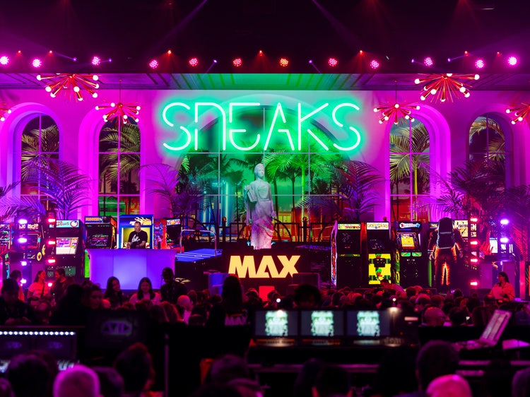 A photo of a stage set with a tropical theme. Ten pinball machines are lined up in front of a man standing at a podium speaking. At the center, front of the stage is MAX spelled out in yellow, all-cap, sans serif type. Writ large among palm trees and purple lighting the word SNEAKS is spelled out in neon lights.