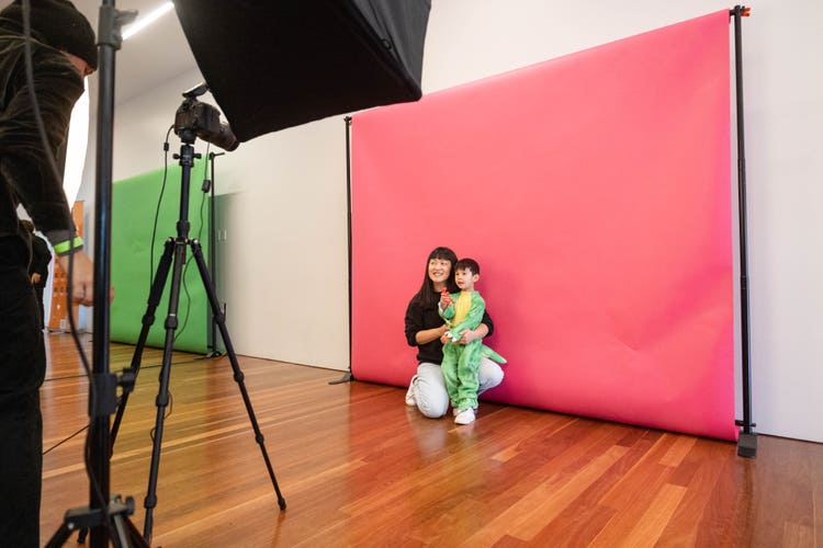 A photograph of a little boy, wearing a green and yellow dinosaur costume, and his mom. He's standing, and she's kneeling with her arms around him, in the background against a pink screen while a photographer in the foreground (using a camera on a tripod) takes their photo.