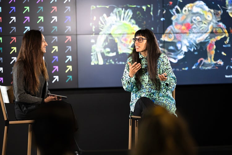 Two white women, both with long dark hair, are seated on stage in conversation, with a large screen divided into two sections. The section on the left is a grid of colorful arrows pointing diagonally upward to the right and on the section on the right is of a colorful abstract artwork.