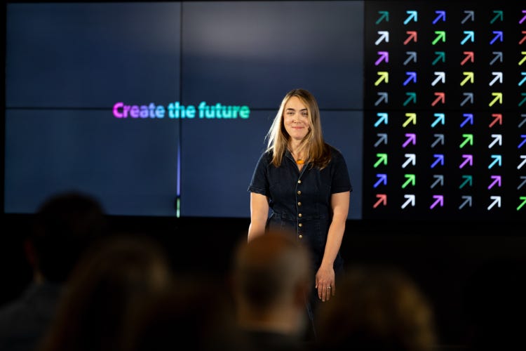 A white woman with shoulder-length blond hair, wearing a denim jumpsuit, stands in front of a large screen divided into two sections. On the left are the words "Create the future" in a pink-to-teal gradient and on the right is a grid of colorful arrows pointing diagonally upward to the right.