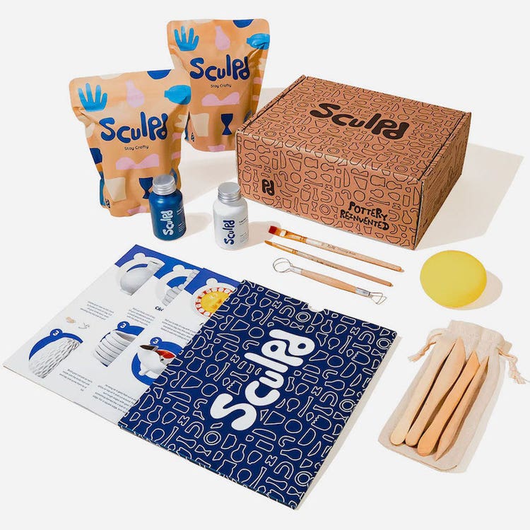 A cardboard box, packages of clay, sculpting tools, bottles of varnish, and a set of instruction manuals—all labeled with Sculpd—arranged on a tabletop.