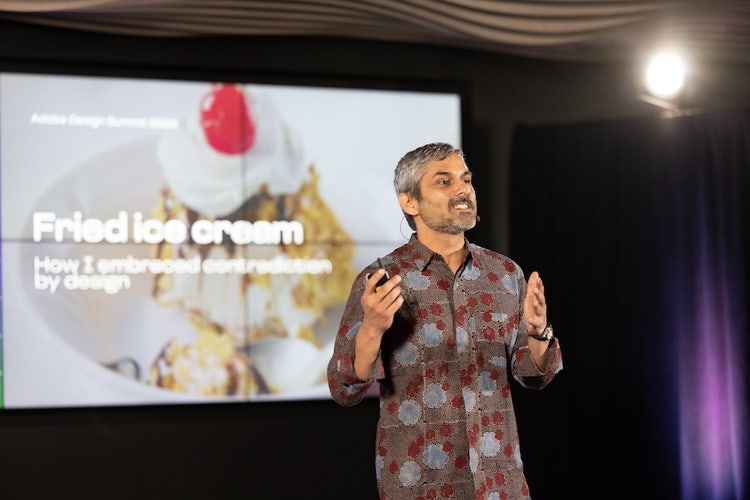 A South Asian man with graying hair and beard, wearing a rust, gray, and brown patterned shirt stands speaking in front of screen that reads "Fried ice cream: How I embraced contradiction by design."