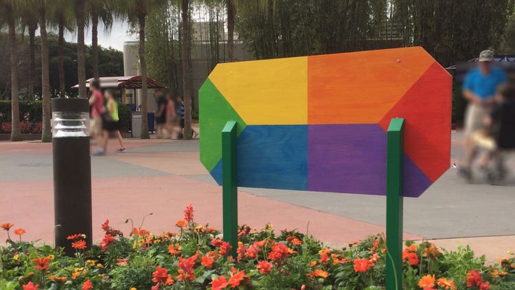 A close-up photograph of the back of rectangular sign in. a flower bed. The sign is divided into six quadrants each painted (clockwise from left): green, yellow, orange, red, indigo, and blue).