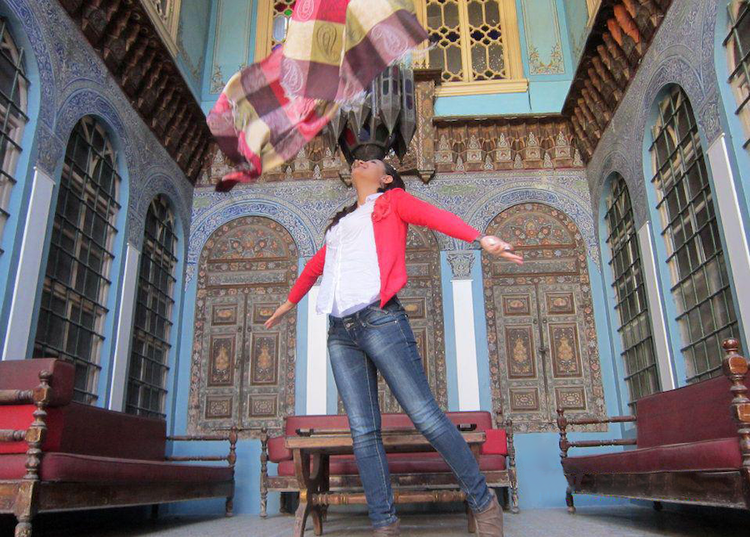 A young woman wearing blue jeans, a white T-shirt, and a red sweater is jumping—with her head thrown back and arms spread wide—and inside a room designed with an Islamic interior. design aesthetic.