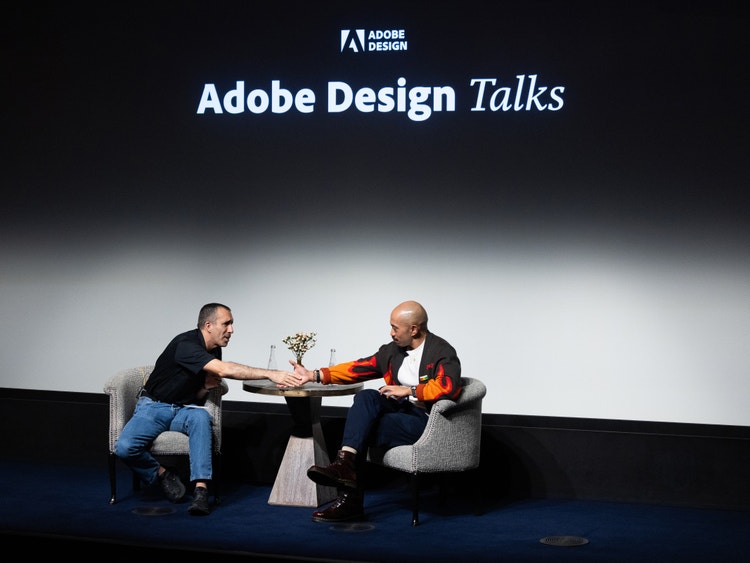 A photograph of two men seated on a stage in front of a digital sign that reads Adobe Design Talks. The man on the left is leaning across a small table to shake the hand of the man on the right.