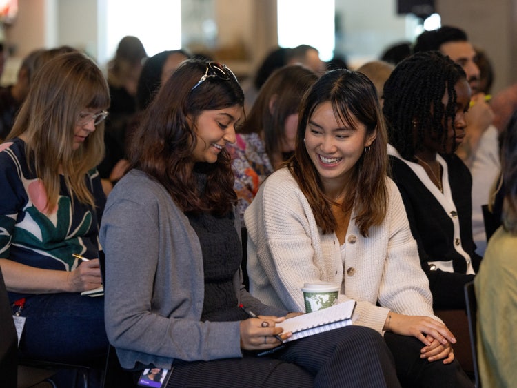 A photograph of a group of people seated in an audience. Two women in the foreground are talking and smiling.