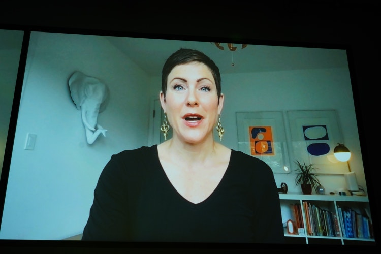 A photograph of a computer screen on which a woman with short dark hair, wearing a black sweater and ornate earrings, is speaking. from a conference call.