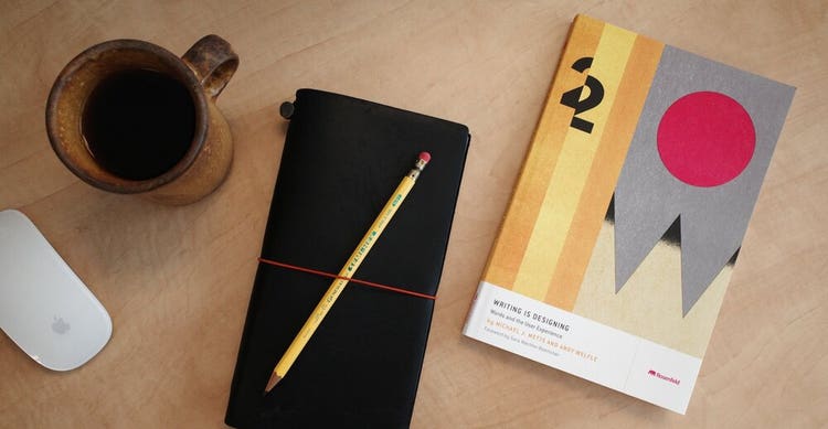 From left to right on a light-colored wood tabletop are a computer mouse, a coffee mug half filled with coffee, a notebook with a pencil secured to it with a rubberband, and a copy of the book "Writing is Designing."