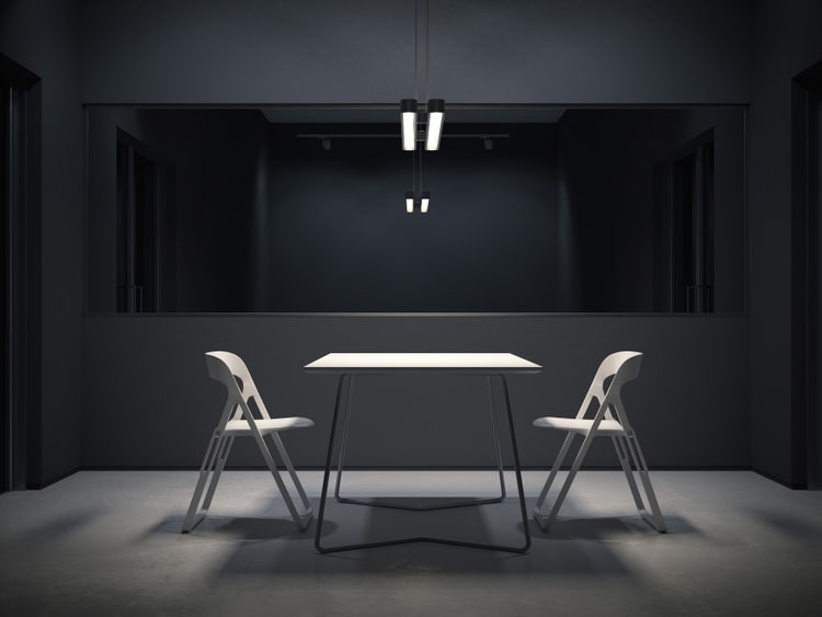 An empty, poorly-lit, interrogation room with a two-sided mirror and in front of it two folding chairs facing each other on either side of a table.