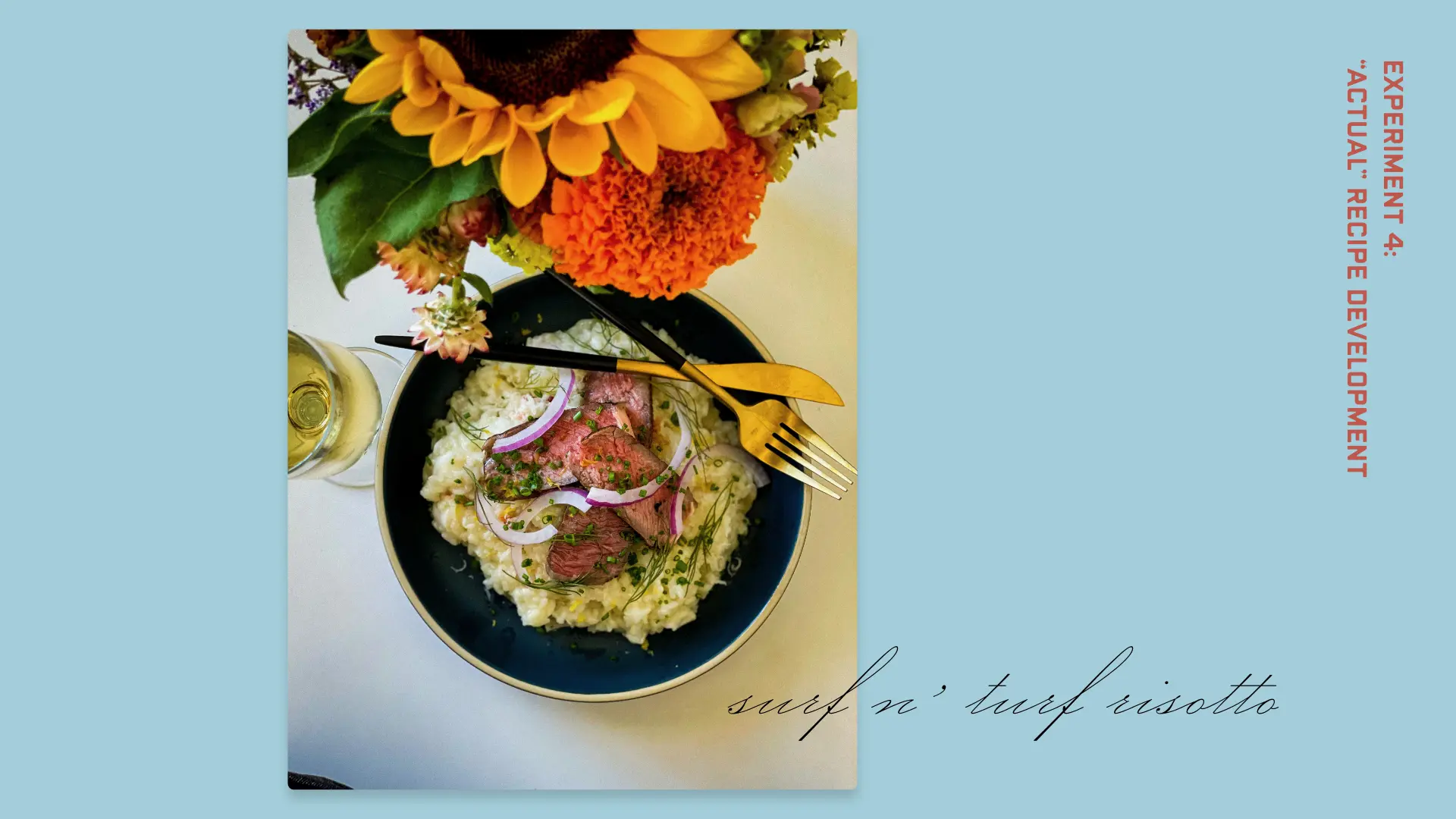A photograph of a place setting on a light blue background. A glass of wine, a flower arrangement, and a knife and fork resting on the edge of a bowl of risotto topped with sliced beef.