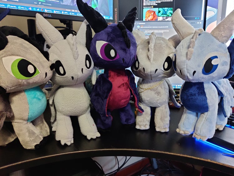 A photograph of a row of five stuffed dragrons "plushies" (each about ten inches tall), in a variety of colors and with a variety of details.