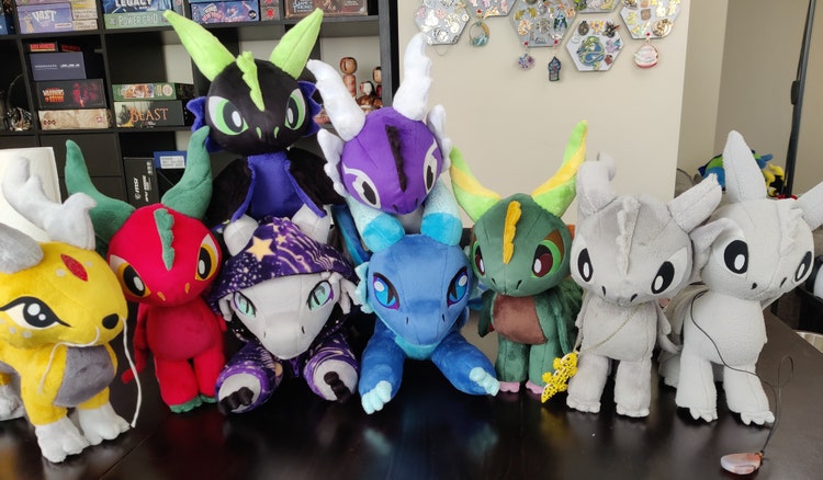 A photograph of a row of nine stuffed dragrons "plushies" (each about ten inches tall), in a variety of colors and with a variety of details.