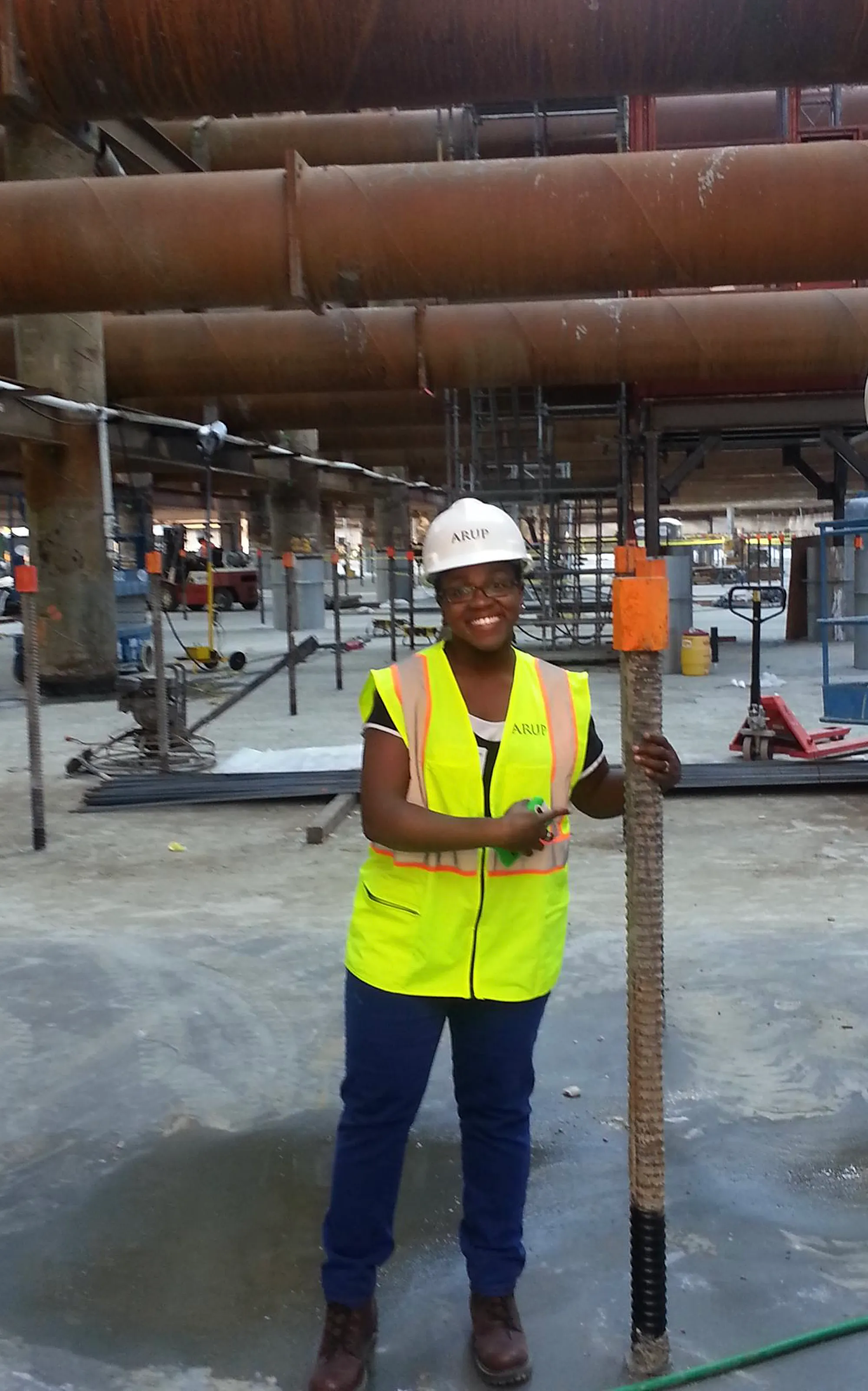 A photograph of a black woman wearing a hard hat, a safety vest, jeans, and work boots poses with rebar in the foreground of a construction site. 