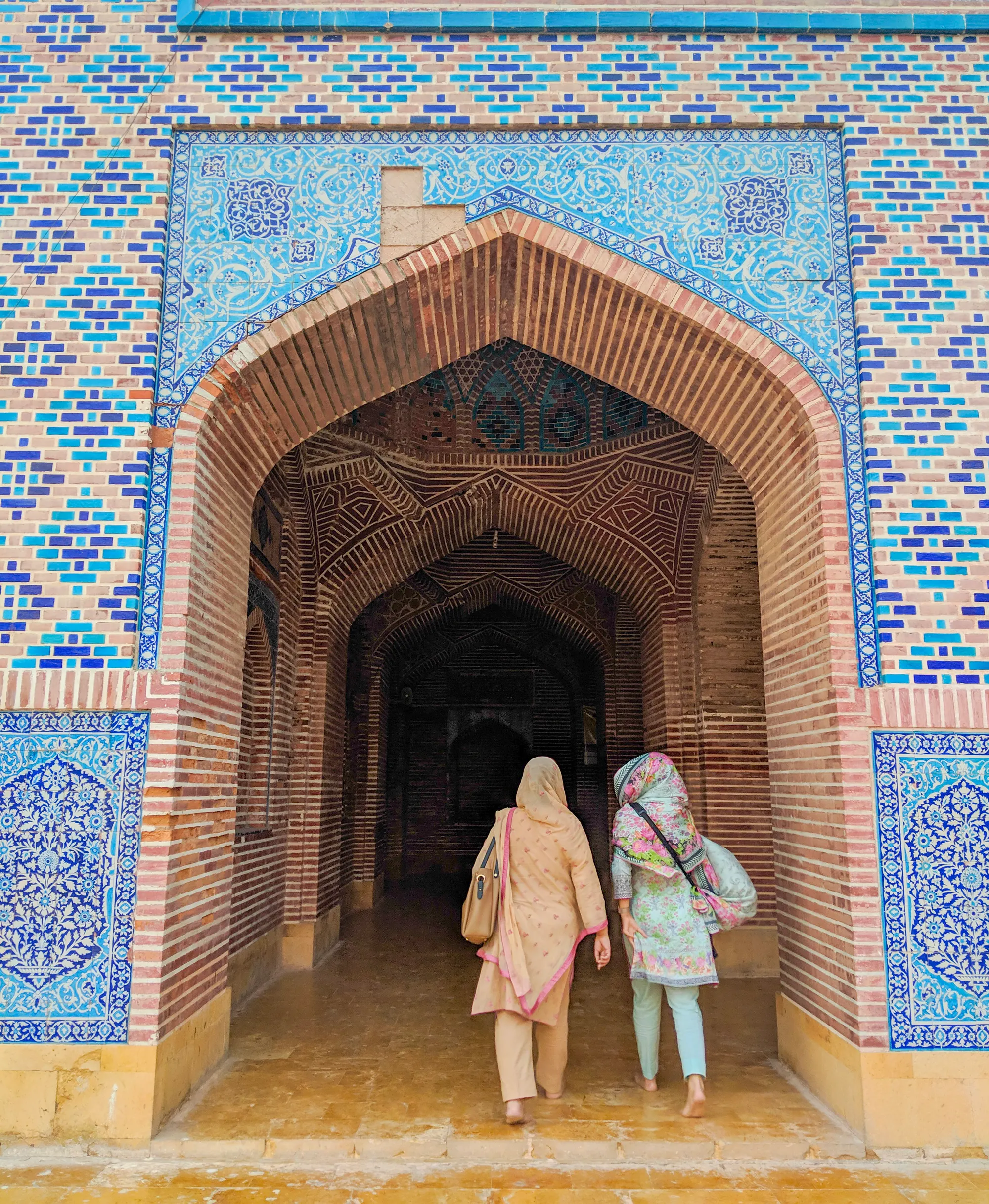 A photograph of two women walking through an arched entrance composed of brick and surrounded by tiles of blue, turquoise, gold, and white set in geometric and floral patterns.
