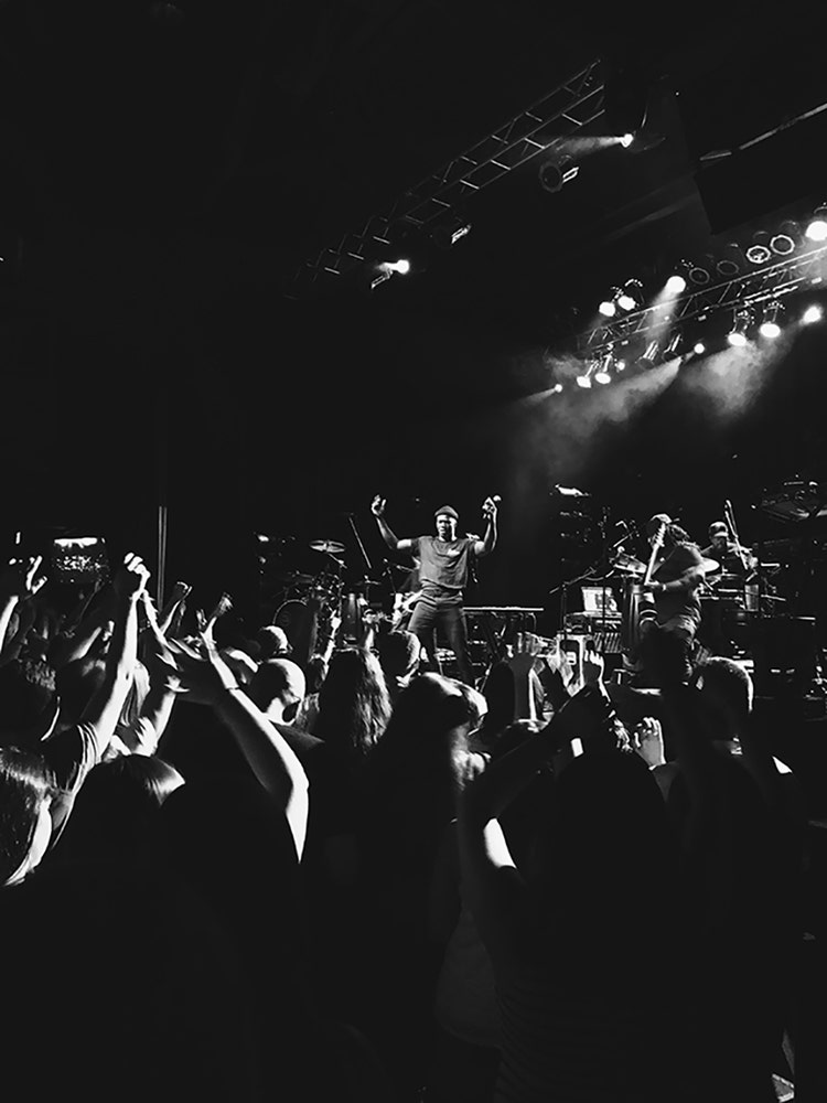 A black-and-white photo of a Black man on a stage wearing a beanie, a T-shirt, and jeans and holding a microphone. On the right side of the stage are two musicians playing string instruments. In the foreground facing him is an audience standing with their hands in the air.