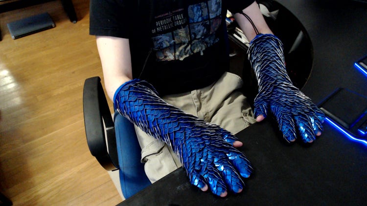 A photograph looking down at man seated in a chair. The view is of his forearms (palms turned down) which are covered fingers to elbows with blue sequin gloves.