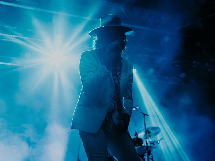 A photograph of a man with shoulder length curly hair wearing a white blazer and a wide flat brim hat. He's holding. a microphone and behind him are a drum set and bright stage lights creating a background of shades of purple and blue.