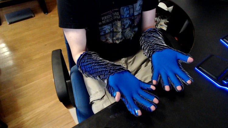 A photograph looking down at man seated in a chair. The view is of his forearms (palms turned up) which are covered from fingers to wrists with blue fabric and from wrists to elbows with blue sequins.