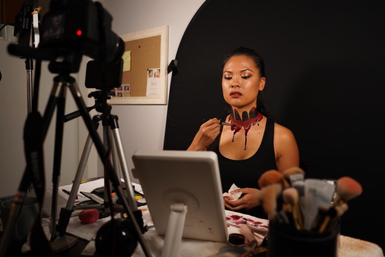 A photograph of a woman seated facing a video camera on a tripod. She's against a black backdrop applying red and black stage makeup to her neck with a brush.