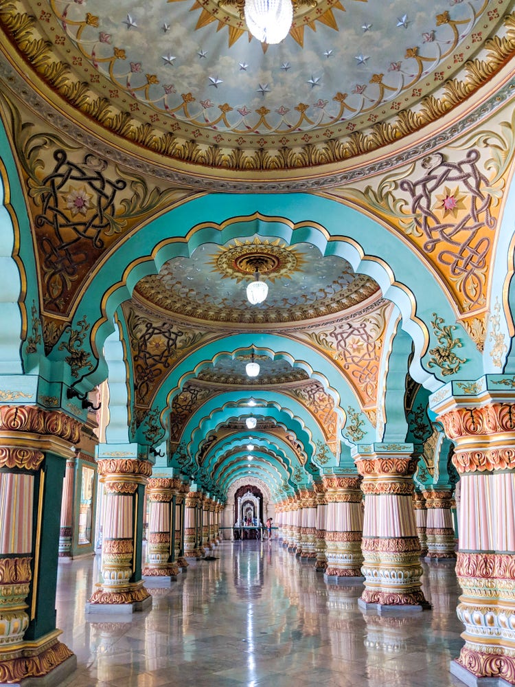A photograph of a great hall of guilded domes, sculptued pillars and archways, and marble floors that form a grand walkway of pink and teal and gold.