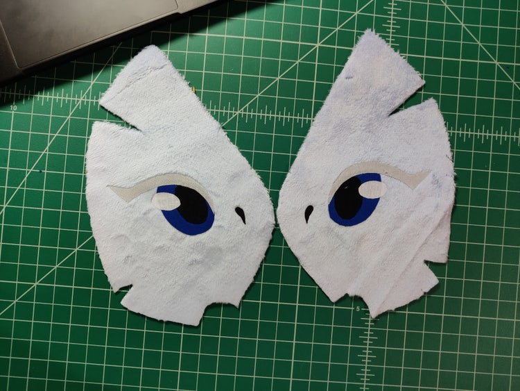 A photograph of two cut fabric pieces that will eventually become the stuffed toy's face are laid out in mirror image (with eye placement) on a green graph-grid cutting board.