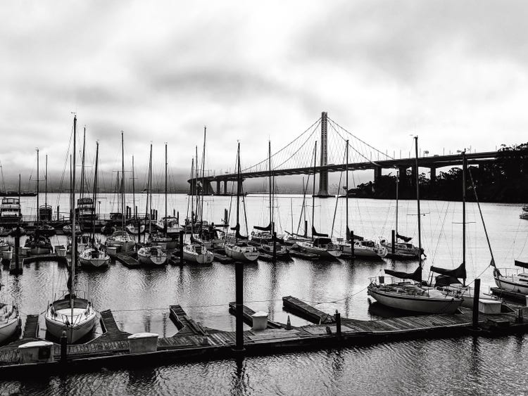 A black-and-white photograph of a marina on a cloudy day. Multiple docked boats are in the foreground and the San Francisco Oakland Bay Bridge is in the background.