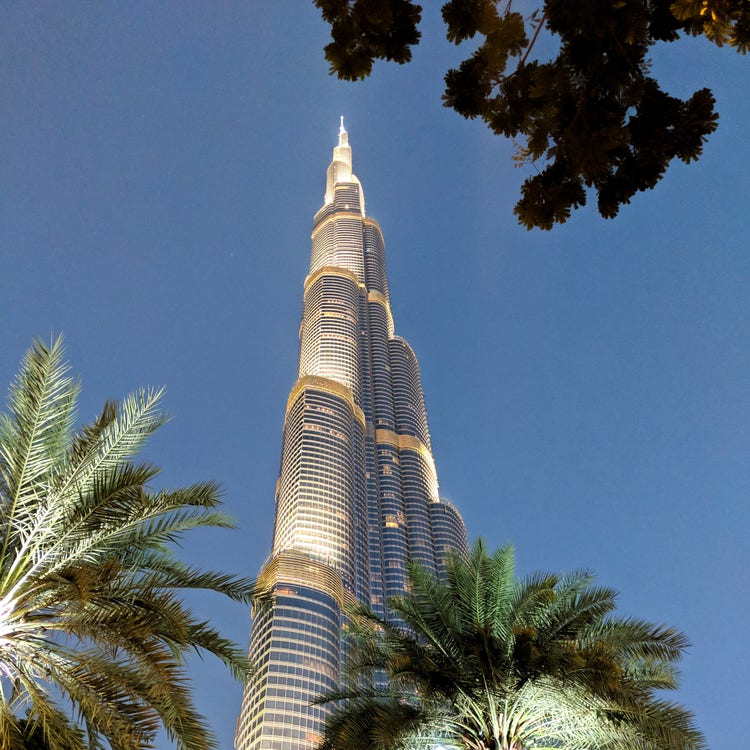 A photograph looking up,  through a foreground of  palm trees, at a majestic golden tower set against a blue sky. 
