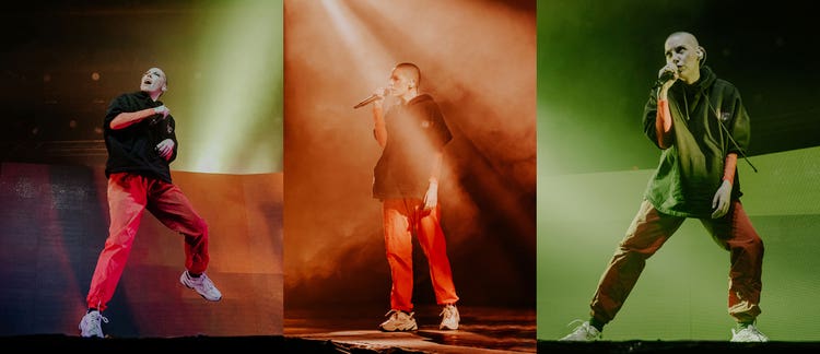 Three photographs of the same white woman with a shaved head. She's wearing a loose dark green pullover hoodie over an orange long-sleeve T-shirt, orange pants, and white sneakers. In the photo on the left, against a green and orange background, she's holding a microphone in her right hand, jumping, and looking up at the ceiling. In the middle photo, against an orange smoke-filled background, she's holding a microphone in her right hand and looking to the right of the stage. In the photo on the right, she's standing in a narrow lunge, against a green background, while facing straight ahead with her eyes focused upward.