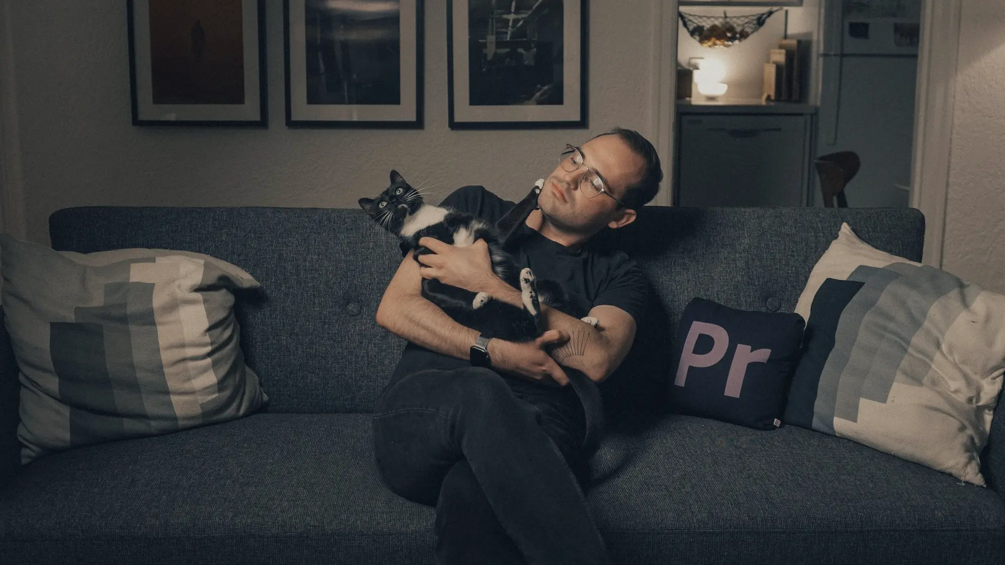 A photograph of a man with glasses sitting on a charcoal grey couch holding a black-and-white cat that's staring at the ceiling.