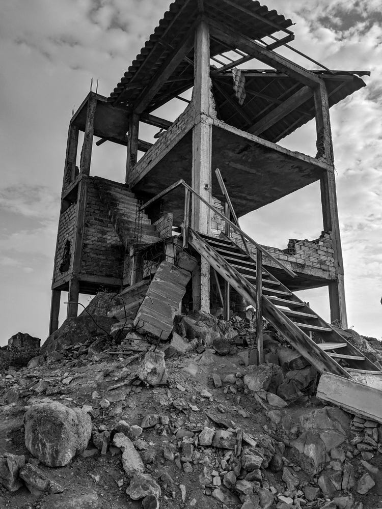 A black-and-white photograph of a crumbling three-story structure  on a rocky hillside. 