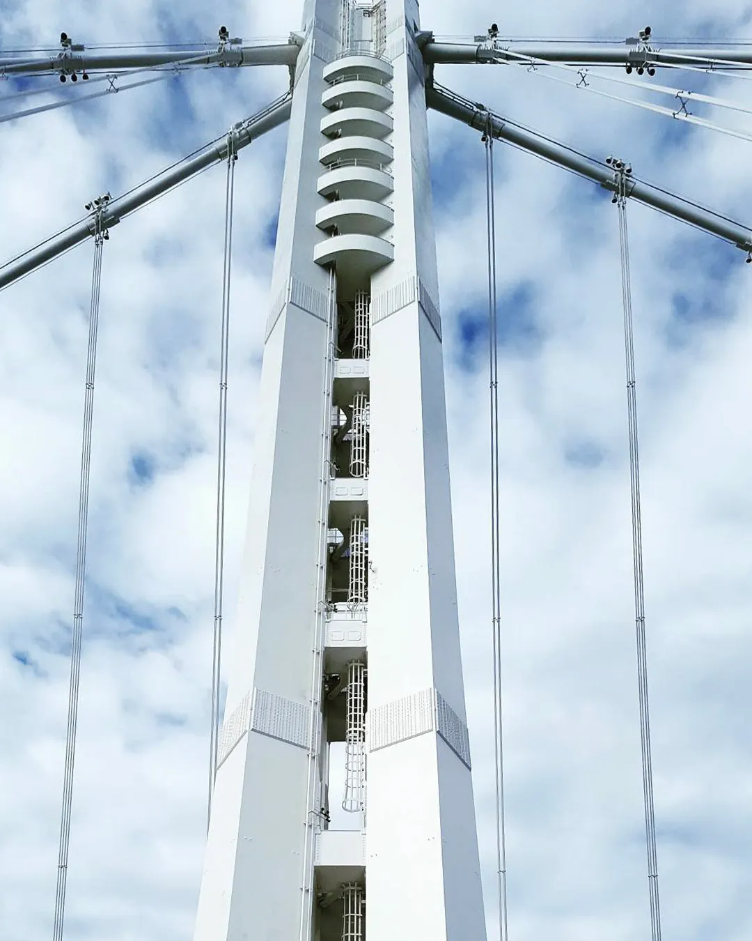 A photograph of a close-up view of a bridge tower and cable structure set against a cloudy blue sky. 