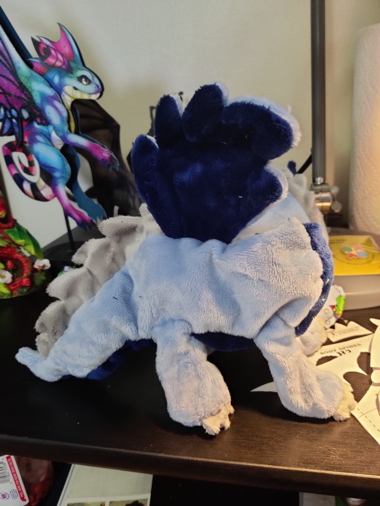 A photograph of a sewn, yet still unstuffed, plush dragon with a purple head, lavendar body, and light grey feet and scales.