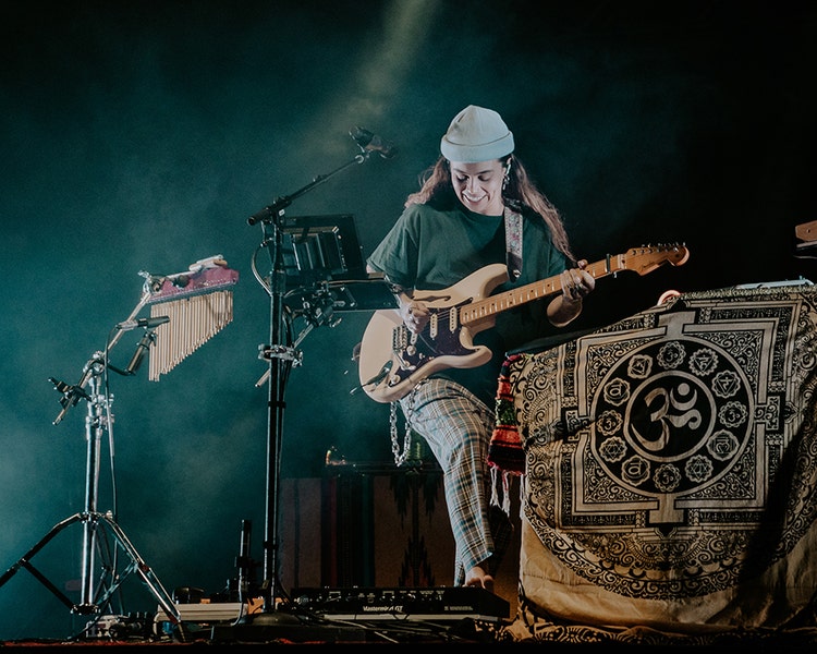 A photograph of a white woman with long brown hair wearing a beanie, a green T-shirt, and plaid pants. She's looking down and smiling while playing a bass guitar and her bare foot is on a mixing board pedal.