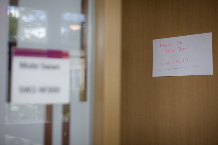 An out-of-focus photo of a conference room window and door. In focus, and taped to the door, is a white index card with a sign hand-written in pink marker that reads, "Premiere Pro Design Jam! Tue 6/11 - Thu 6/13. Please do not erase whiteboard and please leave our snacks. :)"