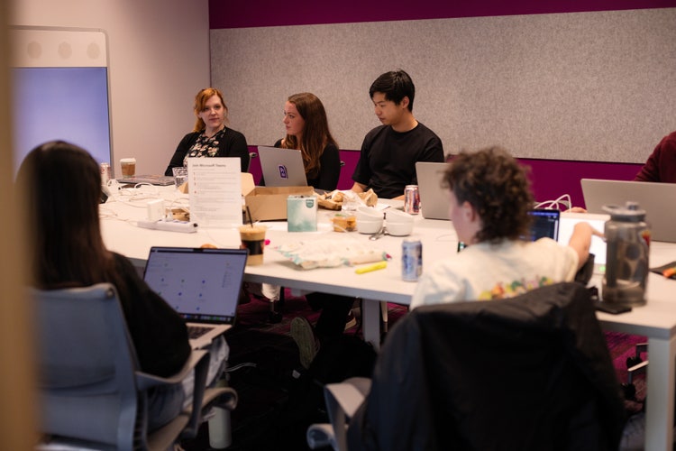 Six men and women seated around a large conference table with their laptops open and facing a large display at the front of the room.