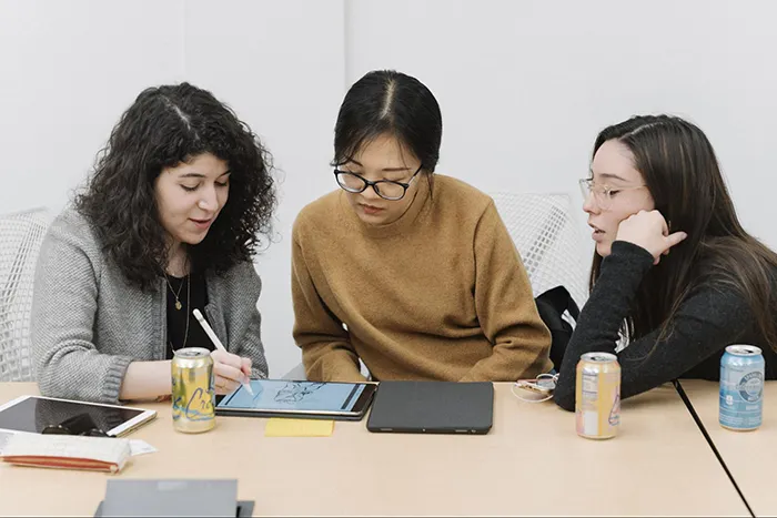 Three women seated together at a conference table are all looking at the same iPad while one of the women draws on it with an Apple Pencil.