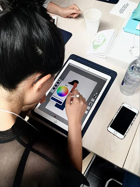 A seated woman with black hair, wearing a sheer black top looks down at her iPad while drawing with an Apple Pencil. On the table are a water bottle, and an iPhone.