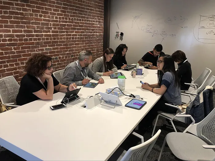 Seven people seated around a conference table drawing on iPads with an Apple Pencils in a room with a brick wall and a dry-erase wall.