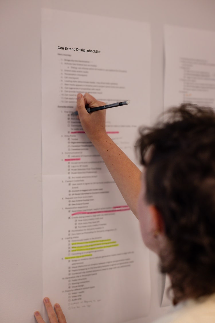 An out-of-focus photo of a person crossing out an item on a sheet of paper, with black text and sections highlighted in pink and yellow, taped to a whiteboard. The only content of the photo that's in focus is a heading at the top of the sheet of paper that reads "Gen Extend Design Checklist."