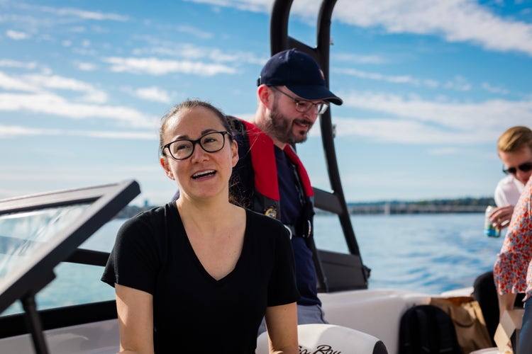 Four men and women on a boat, with one white woman wearing glasses and a black T-shirt directly facing the camera, against an out of focus view of the water and the horizon behind them.